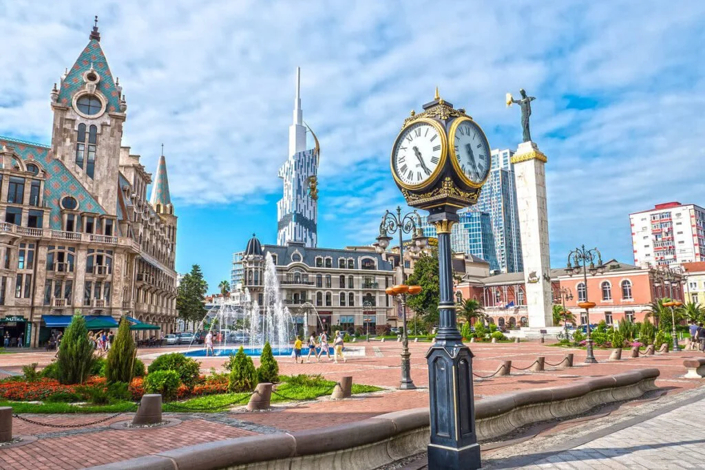 City square with a fountain, a decorative clock, a statue on a tall pedestal, and a mix of historic and modern buildings in the background, under a partly cloudy sky.