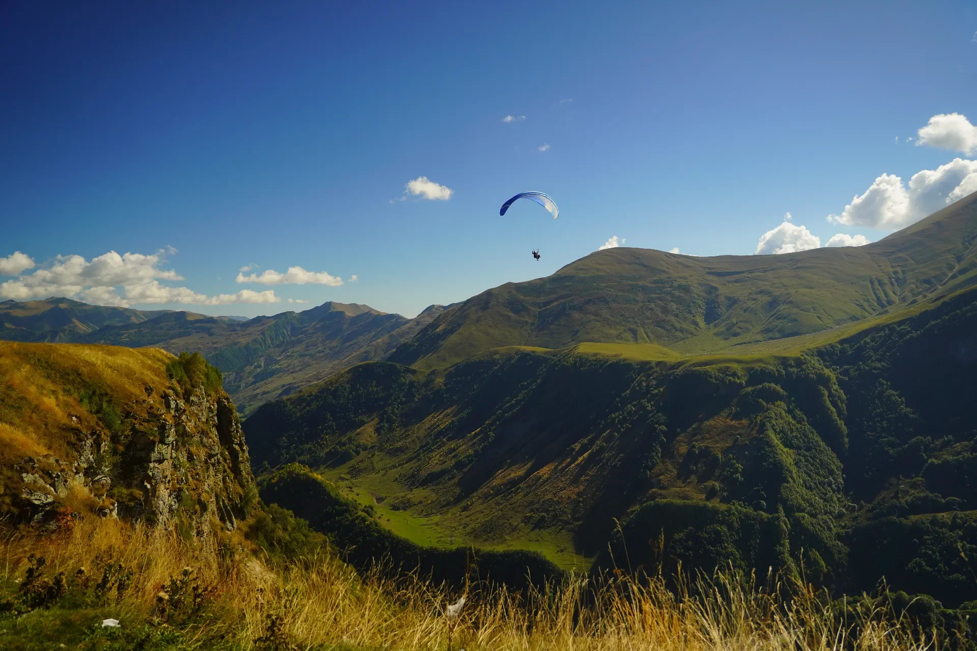 A paraglider flying over a lush mountainous landscape with green hills and valleys, under a bright blue sky with scattered clouds.