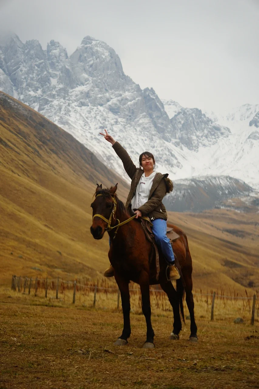 A woman riding a horse in a mountainous landscape with snow-covered peaks in the background, gesturing with her right hand.
