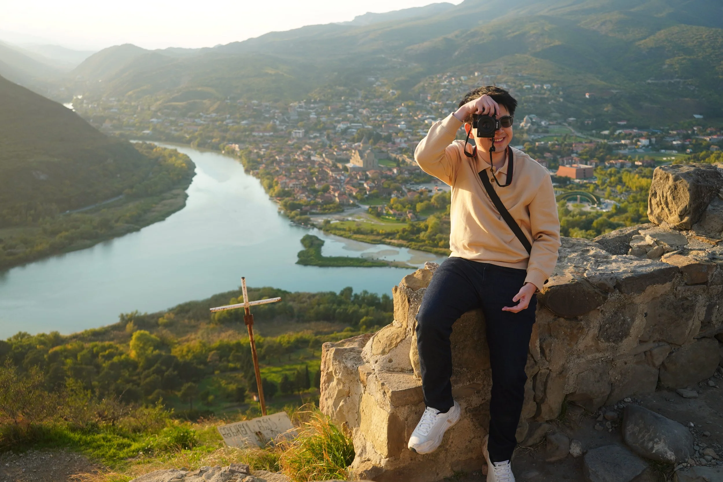A young man taking a photo with a camera while sitting on a stone wall on a hill overlooking a river and a small town in a valley surrounded by mountains.