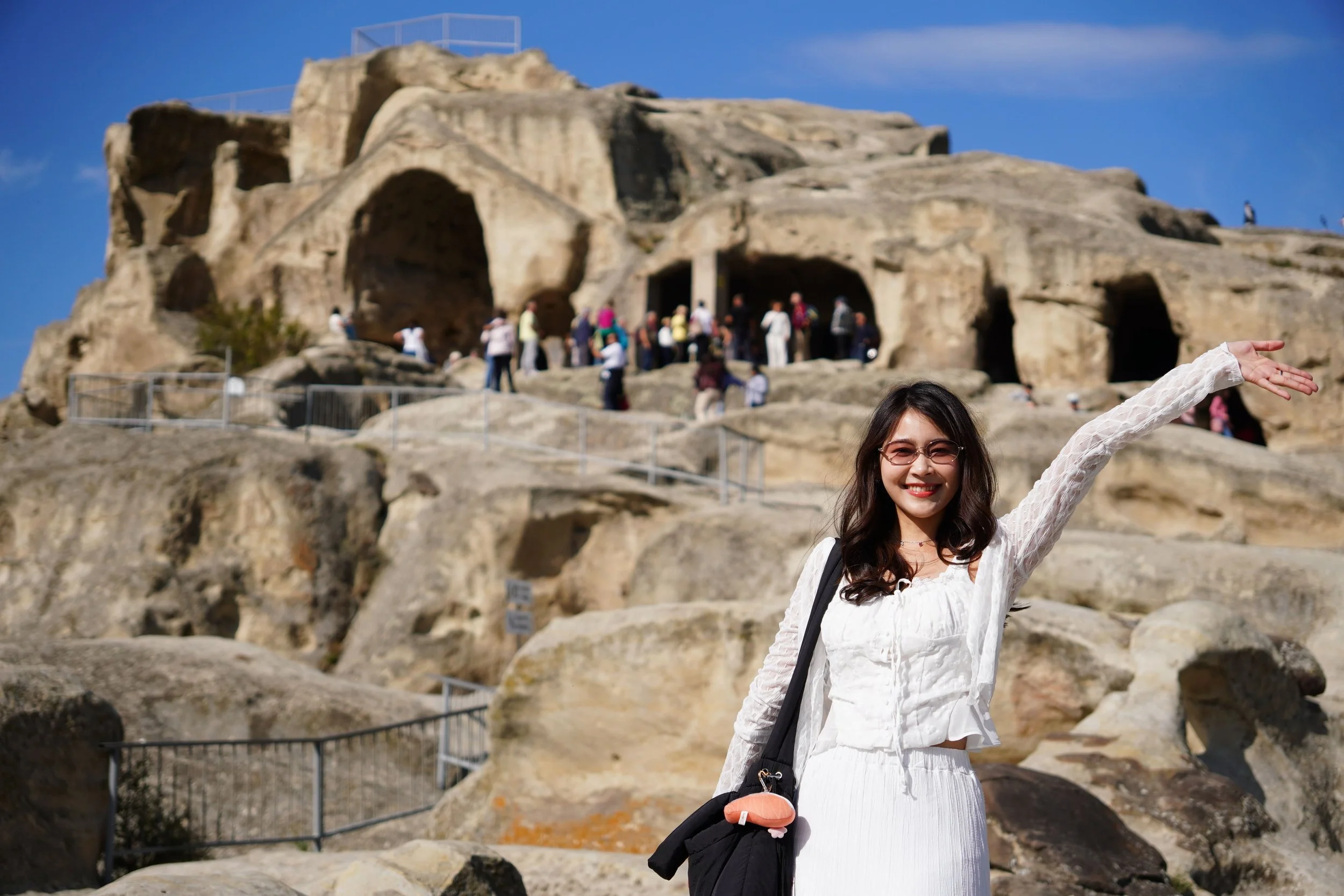 A young woman in a white dress and sunglasses smiling and pointing to the right, standing in front of ancient rock formations and caves with tourists exploring in the background.