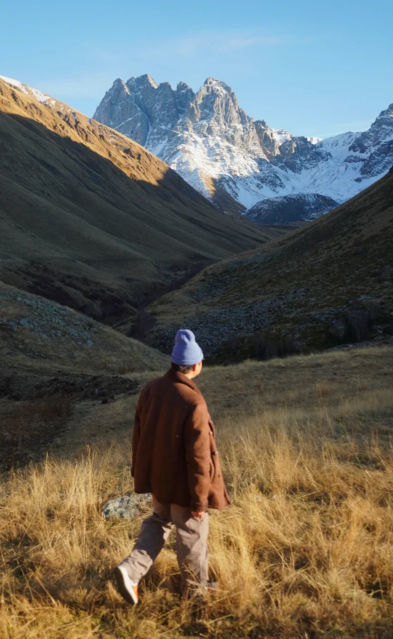 A person wearing a brown jacket and a blue beanie walking through a grassy valley with mountains in the background, some snow on the peaks, under a clear blue sky.