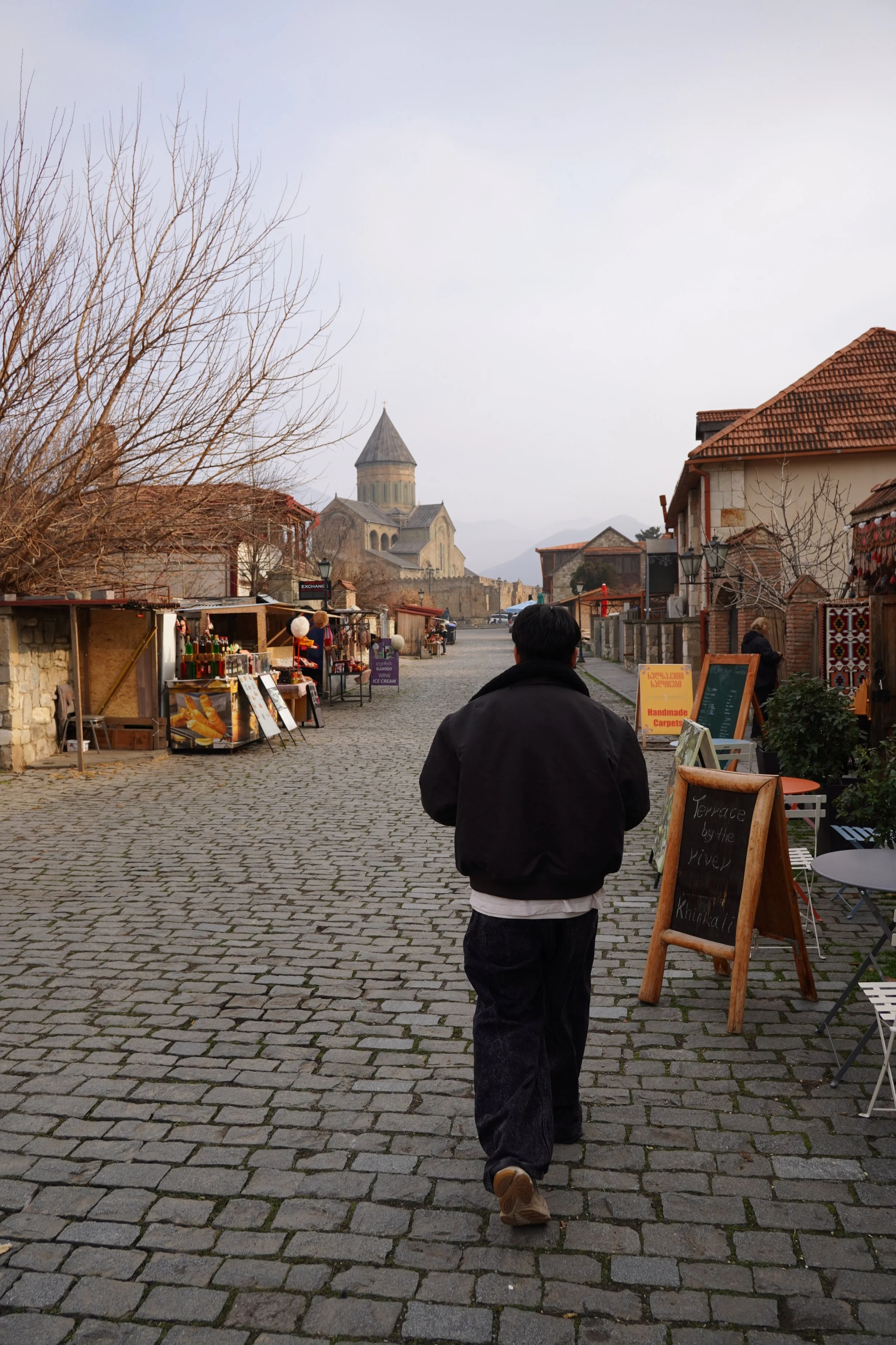 A man walking on a cobblestone street in a European town, with market stalls on the side, leafless trees, and a church in the background.