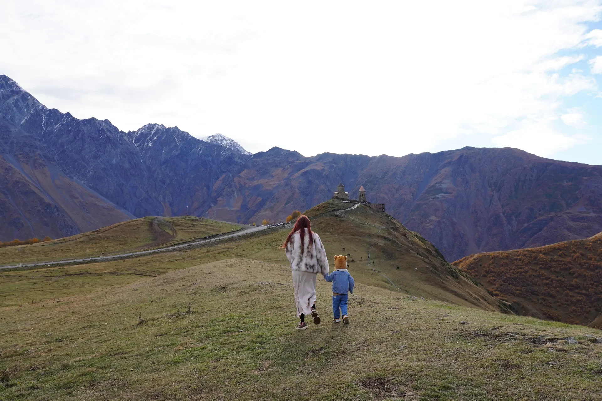 A woman with long red hair and a child with a teddy bear hat walking in a grassy hilly landscape towards a castle on a hill, with mountain ranges in the background.
