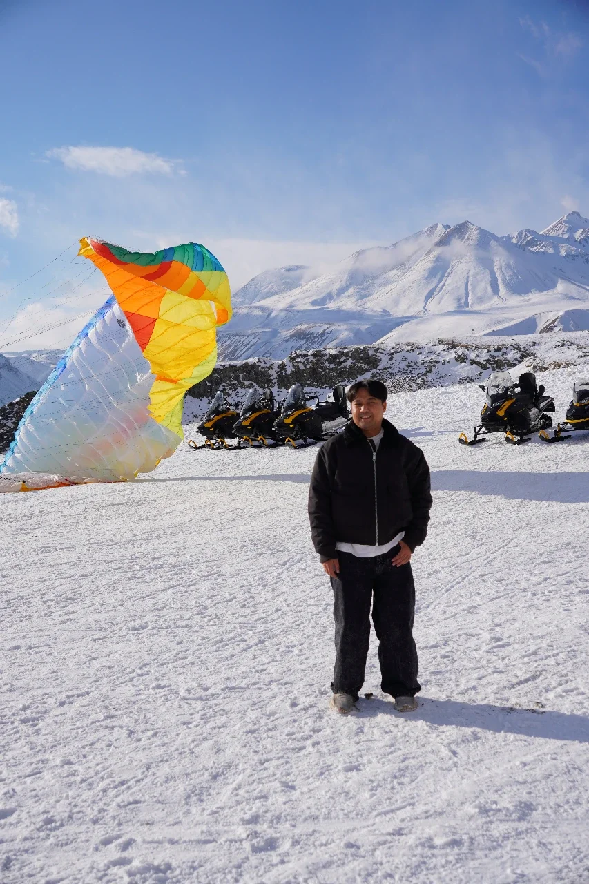 A person standing on snow with a colorful kite and snowmobiles in the background, mountains and blue sky visible.