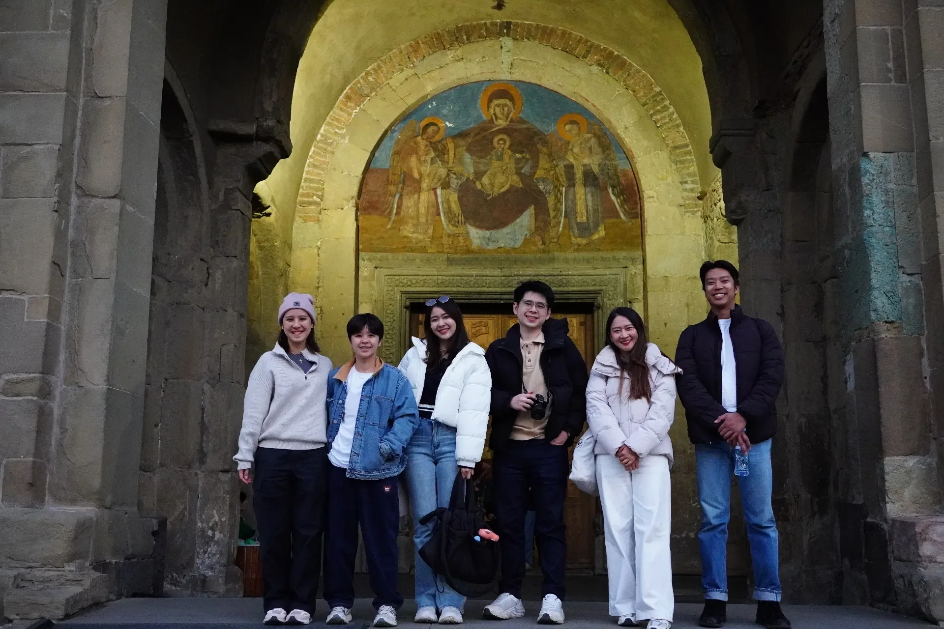 A group of six young people standing together inside a historic church or cathedral, with a religious fresco above the door behind them.