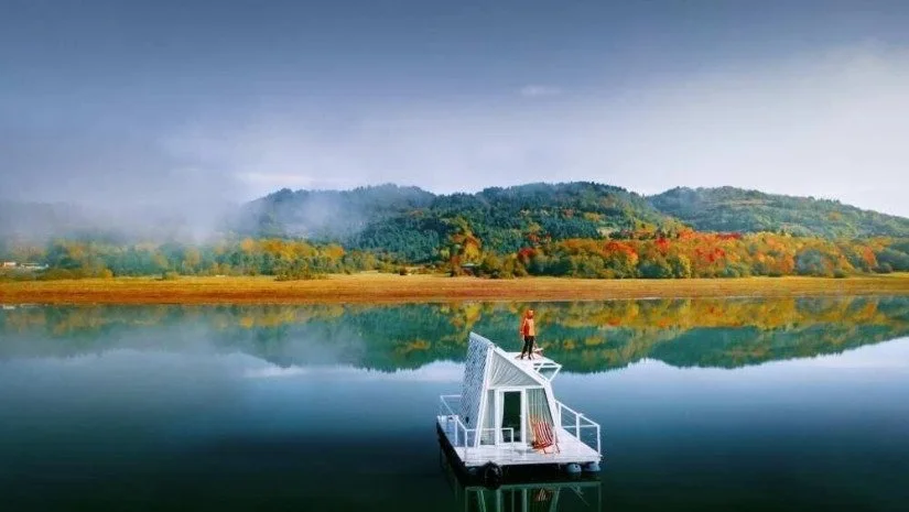 A person standing on a small white houseboat in a calm lake surrounded by mountains with trees showing fall colors.
