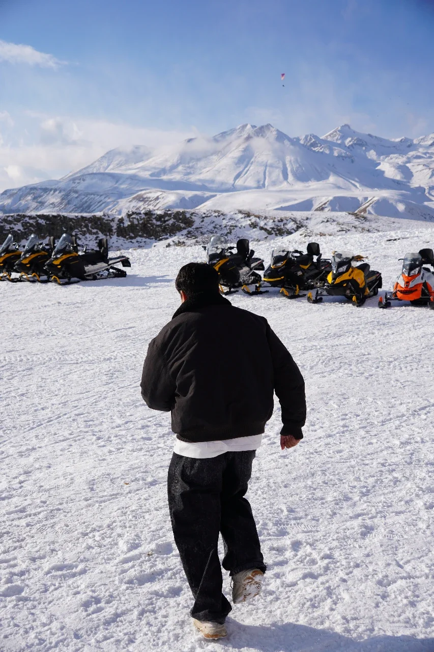 A person walking on snow with snowmobiles parked in the background and snow-covered mountains in the distance under a partly cloudy sky.