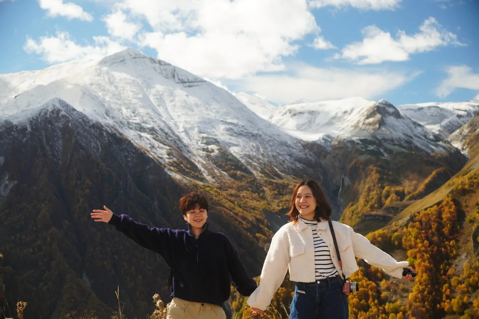 A woman and a boy happily holding hands in a mountainous landscape with snow-capped peaks and colorful autumn trees.