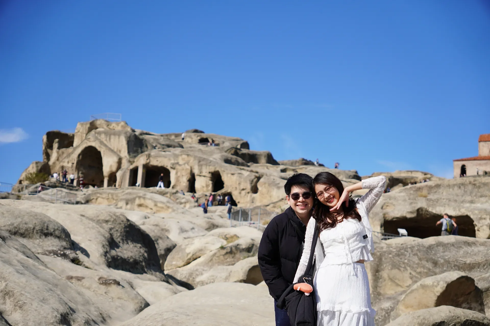 A happy young couple standing in front of ancient cave dwellings carved into rock, with other visitors exploring in the background under a clear blue sky.