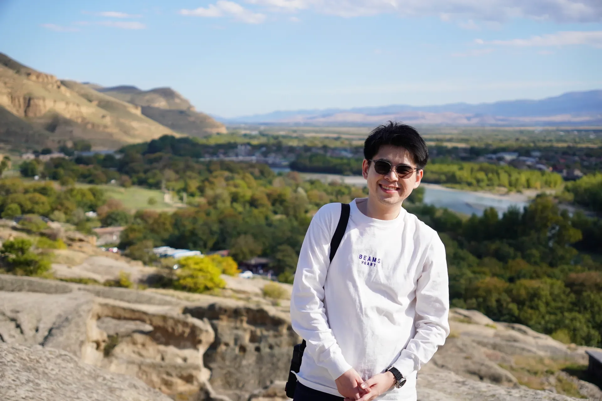 A smiling young man wearing sunglasses and a white long-sleeve shirt standing outdoors on a rocky surface with a scenic view of mountains, green trees, and a river in the background.