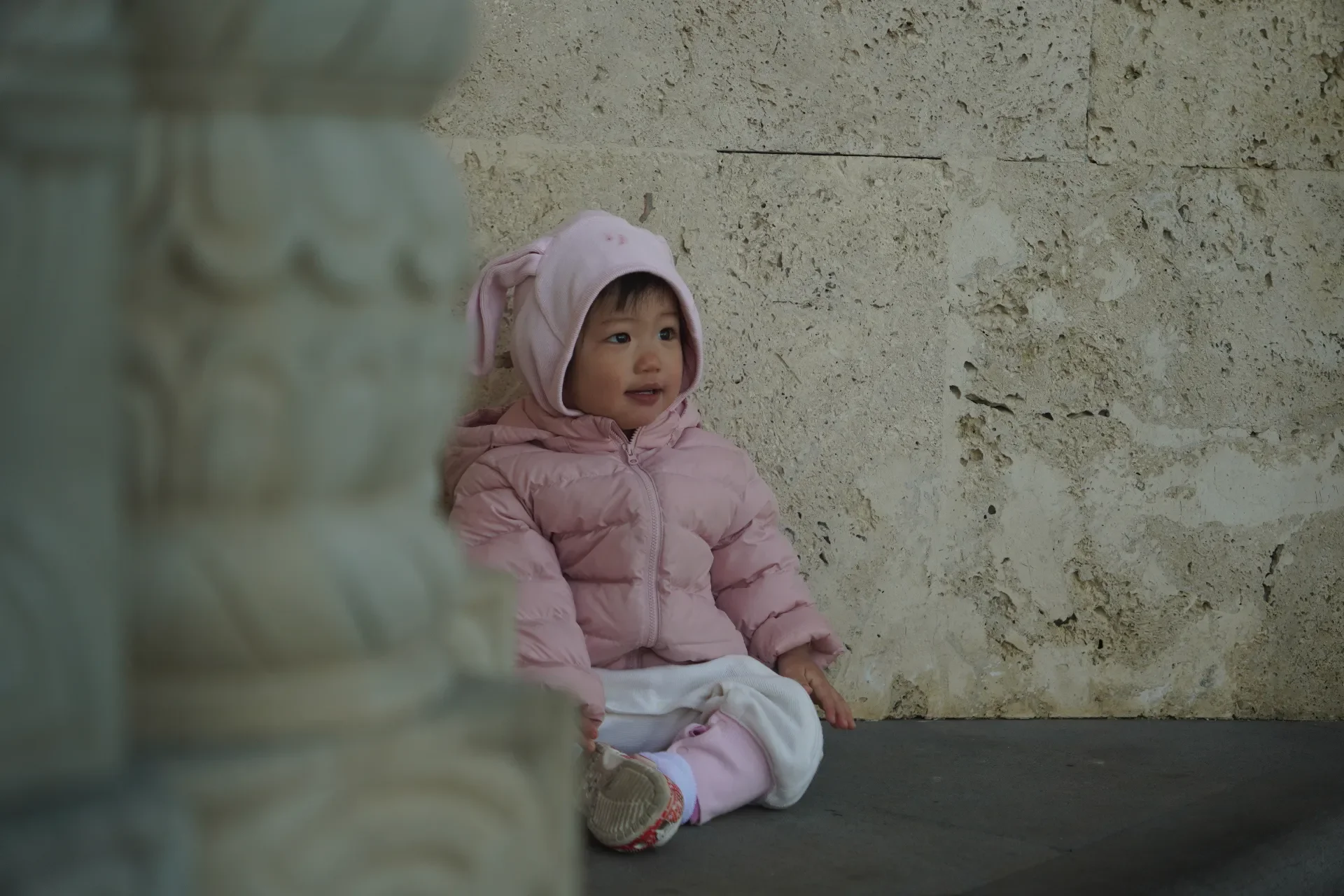 A young child sitting on the ground against a stone wall, wearing a pink jacket, a hood with bunny ears, and white clothes.