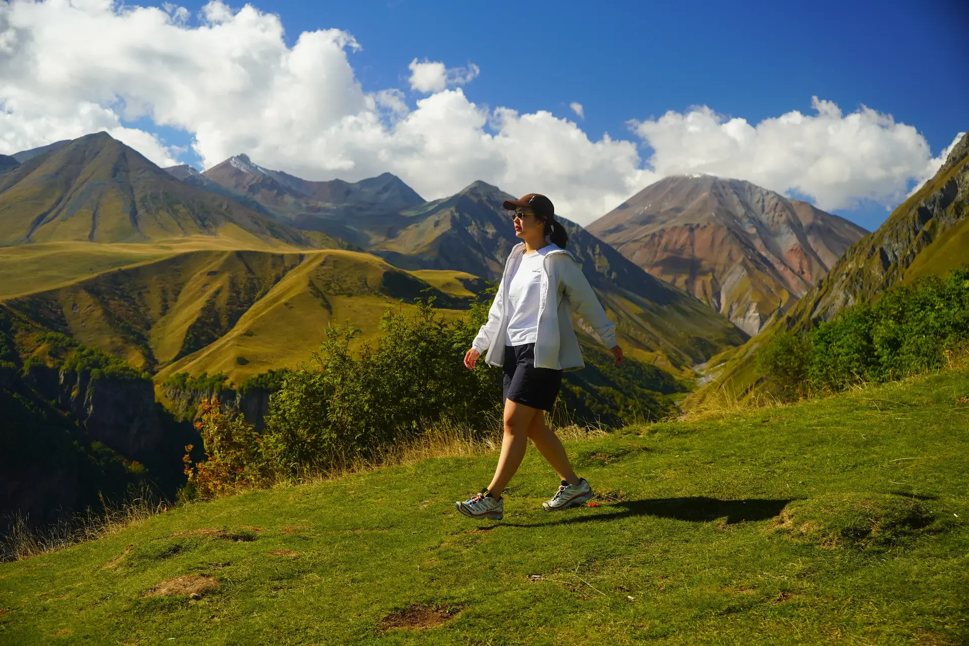 A woman hiking in a mountainous landscape with green hills and rocky peaks under a partly cloudy sky.