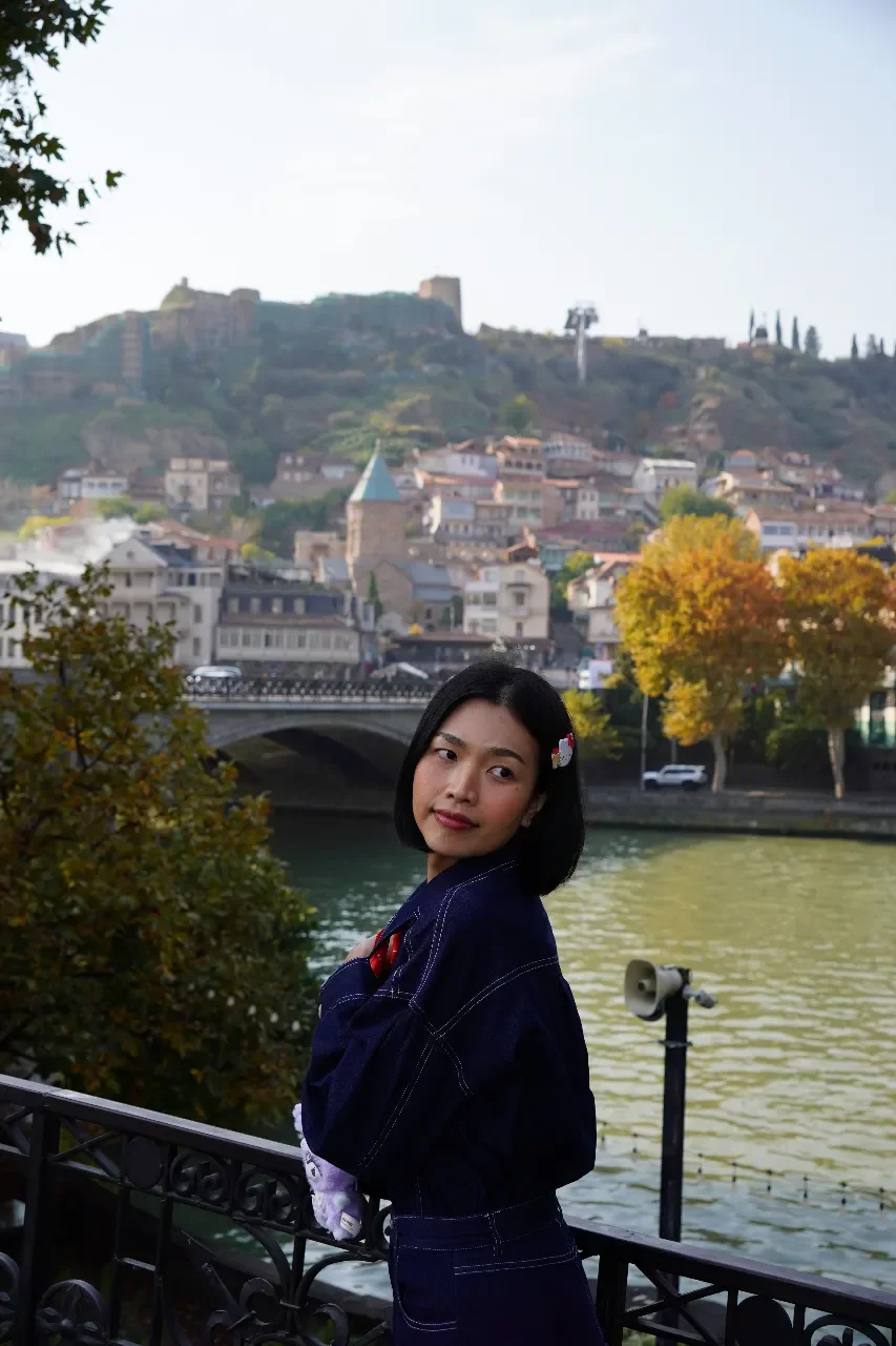 A young woman with short black hair and a white flower hair clip standing near a river, with a historic European-style town and hillside in the background.