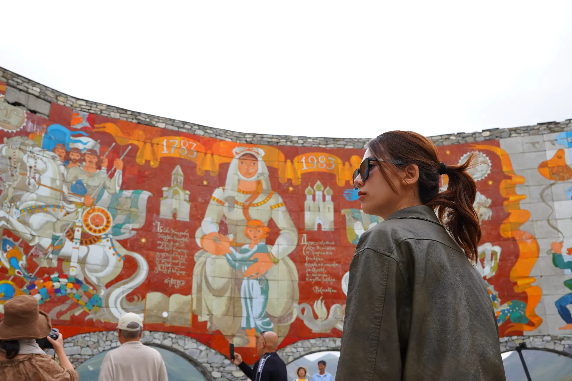 A woman with brown hair tied in a ponytail, wearing sunglasses and a dark jacket, stands in front of a colorful mural depicting historical and mythological figures with the years 1783 and 1983, and other people are taking photos nearby.