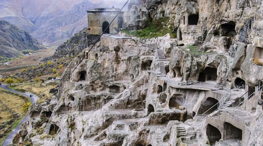 Ancient rock-cut cave dwellings and tunnels carved into a mountainside with staircases and a pathway, overlooking a scenic valley with a winding road.