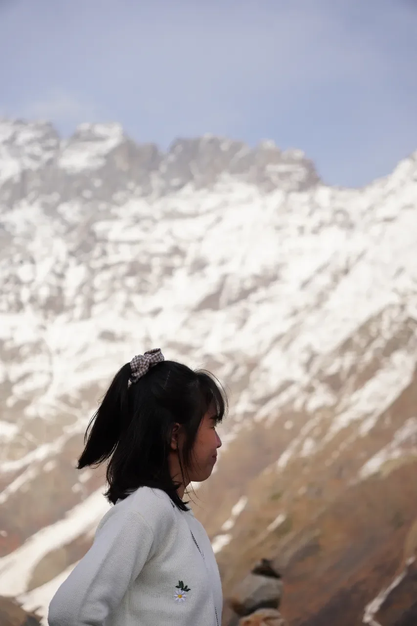 A young girl with dark hair in a ponytail secured by a checkered hair tie, wearing a white sweater with small embroidered flowers, standing outdoors against a rugged, snow-capped mountain landscape.