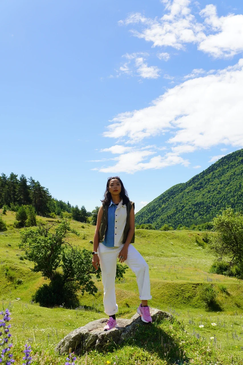 A woman standing outdoors on a large rock in a green valley with rolling hills and a bright blue sky with some clouds.