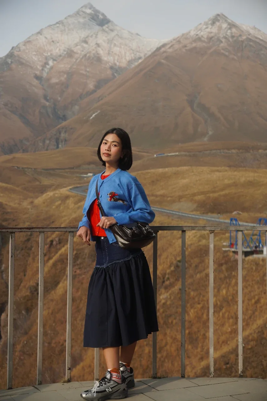 A woman standing outdoors on a balcony with mountain scenery in the background, dressed in casual clothing including a blue jacket, red shirt, black skirt, and sneakers.