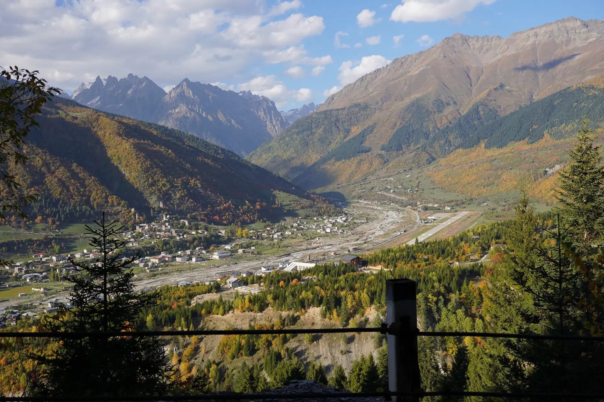 Mountain valley landscape with colorful autumn trees, a small town, and high mountain peaks in the distance under a partly cloudy sky.