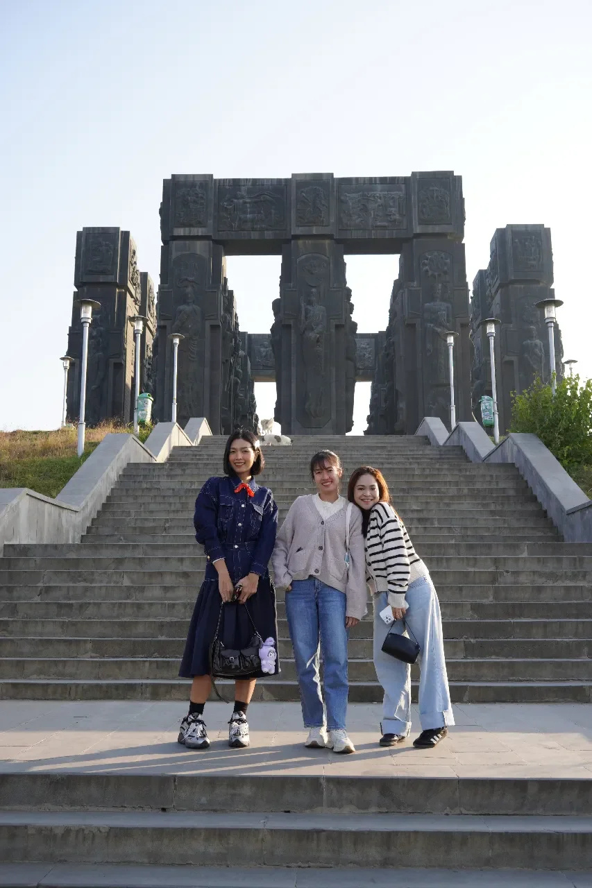 Three young women smiling and posing on steps in front of a large monument with carved figures and open rectangular gaps, during daylight.