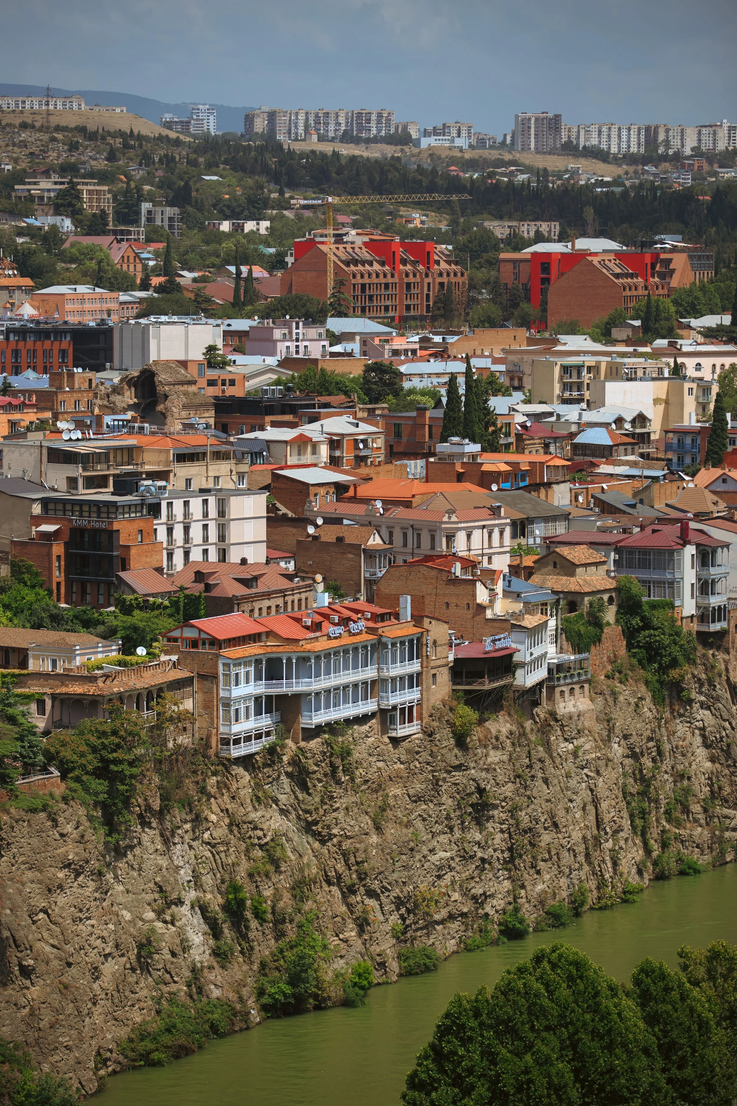 Cityscape with houses and apartment buildings on a hillside above a river, with mountains in the background.