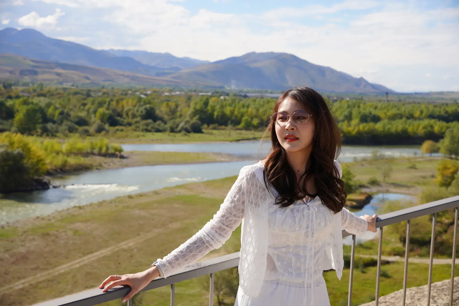 A woman with long dark hair, wearing glasses and a white lace dress, standing on a balcony overlooking a river with mountains in the background during daytime.