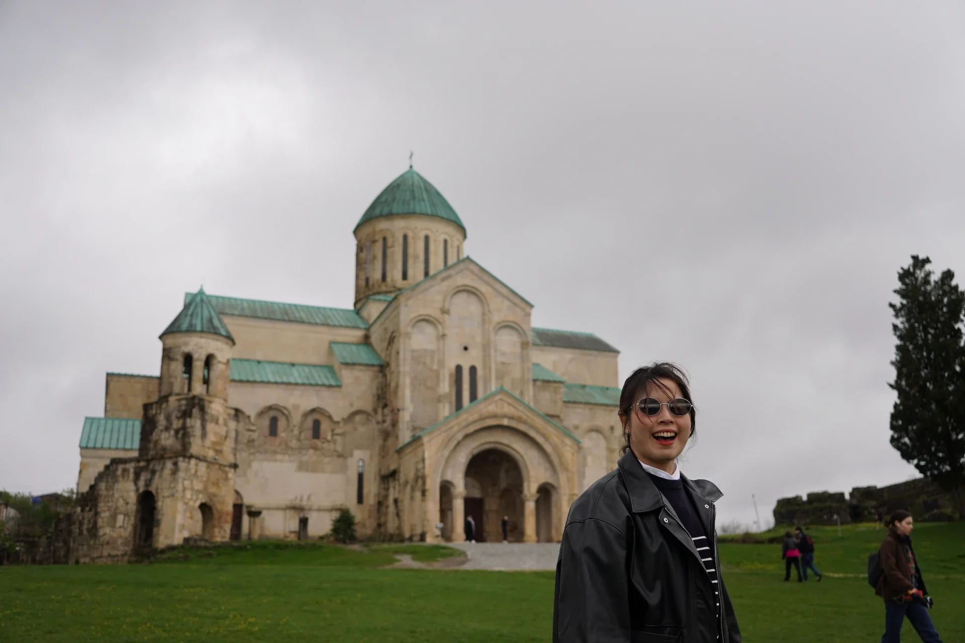 A woman with dark hair wearing sunglasses and a black leather jacket smiling outside a historic stone church with green-roofed towers under a cloudy sky.