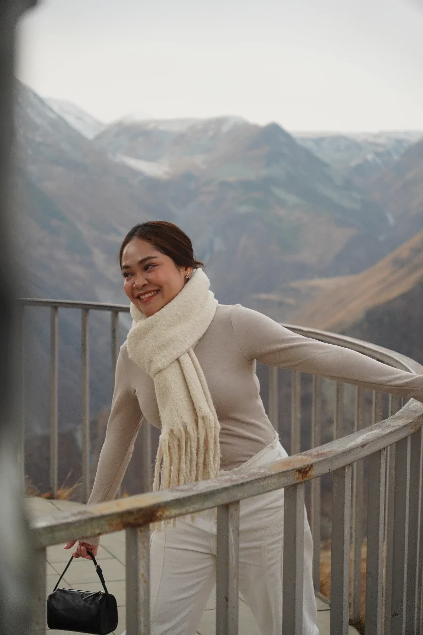 A smiling woman with brown hair pulled back, wearing a cream sweater, white pants, and a large cream scarf, standing on a winding metal staircase outdoors with mountains in the background.
