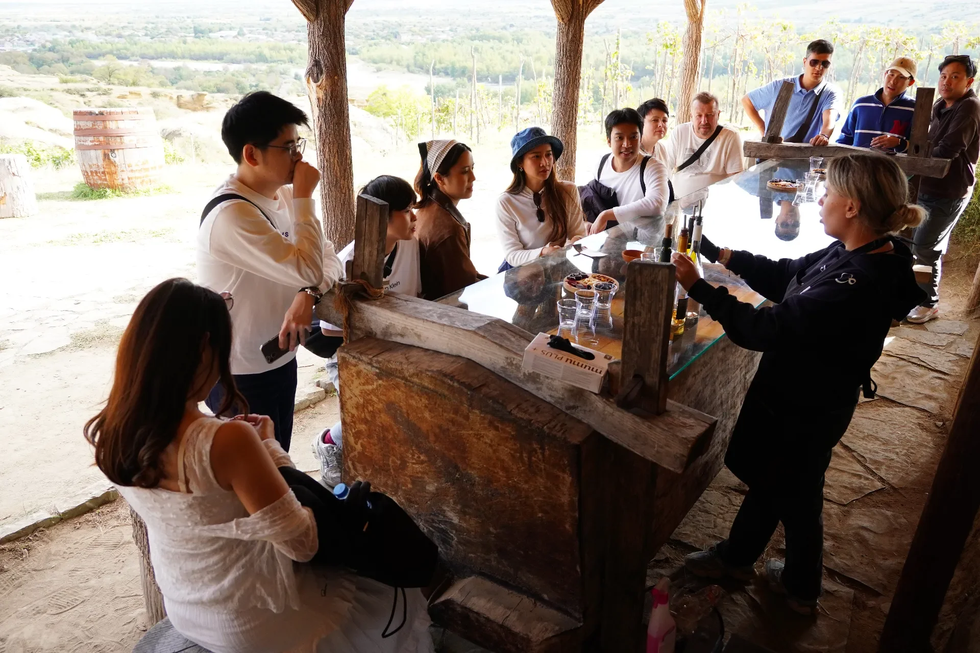 Group of people sitting and standing around a rustic outdoor bar, with drinks and snacks, under a wooden shelter with open sides and trees in the background.