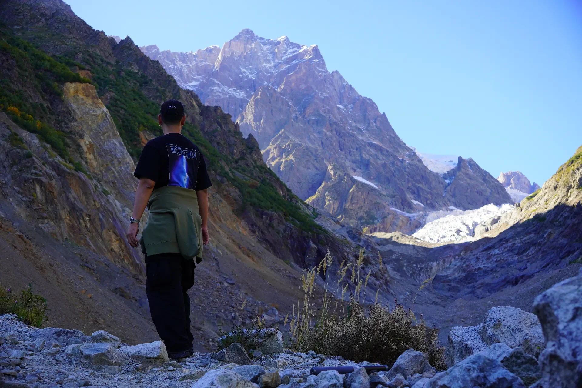A person hiking on a rocky mountain trail with snow-capped peaks and rugged mountains in the background during daytime.