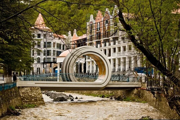 A bridge over a river with uniquely designed circular artwork in the center, surrounded by tall, historic-style buildings and trees with green leaves.