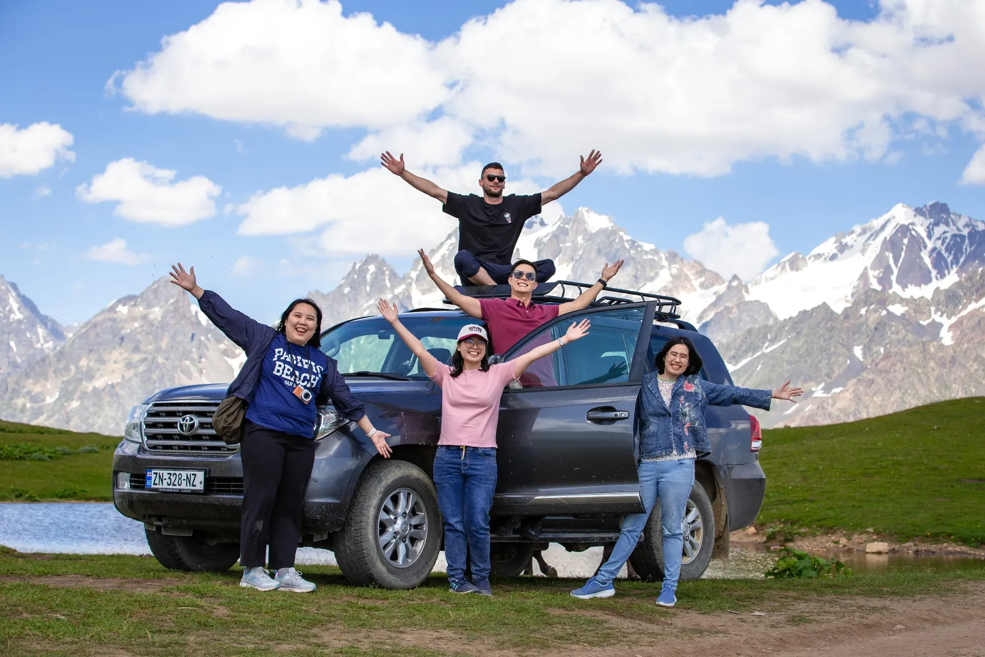 Group of five friends enjoying a day outdoors with a snowy mountain backdrop, standing around and on a dark gray SUV, with three standing on the grass and two sitting on the roof rack, all smiling with arms raised in celebration.