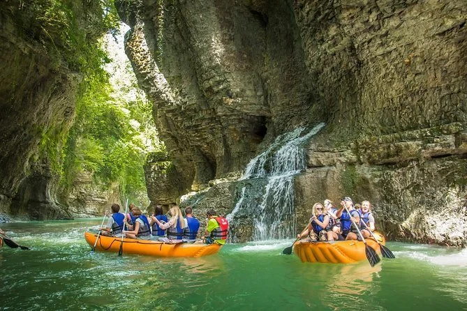 Group of people in two bright orange kayaks floating on a river surrounded by high rocky cliffs and lush green trees, with a small waterfall cascading down the cliffs.
