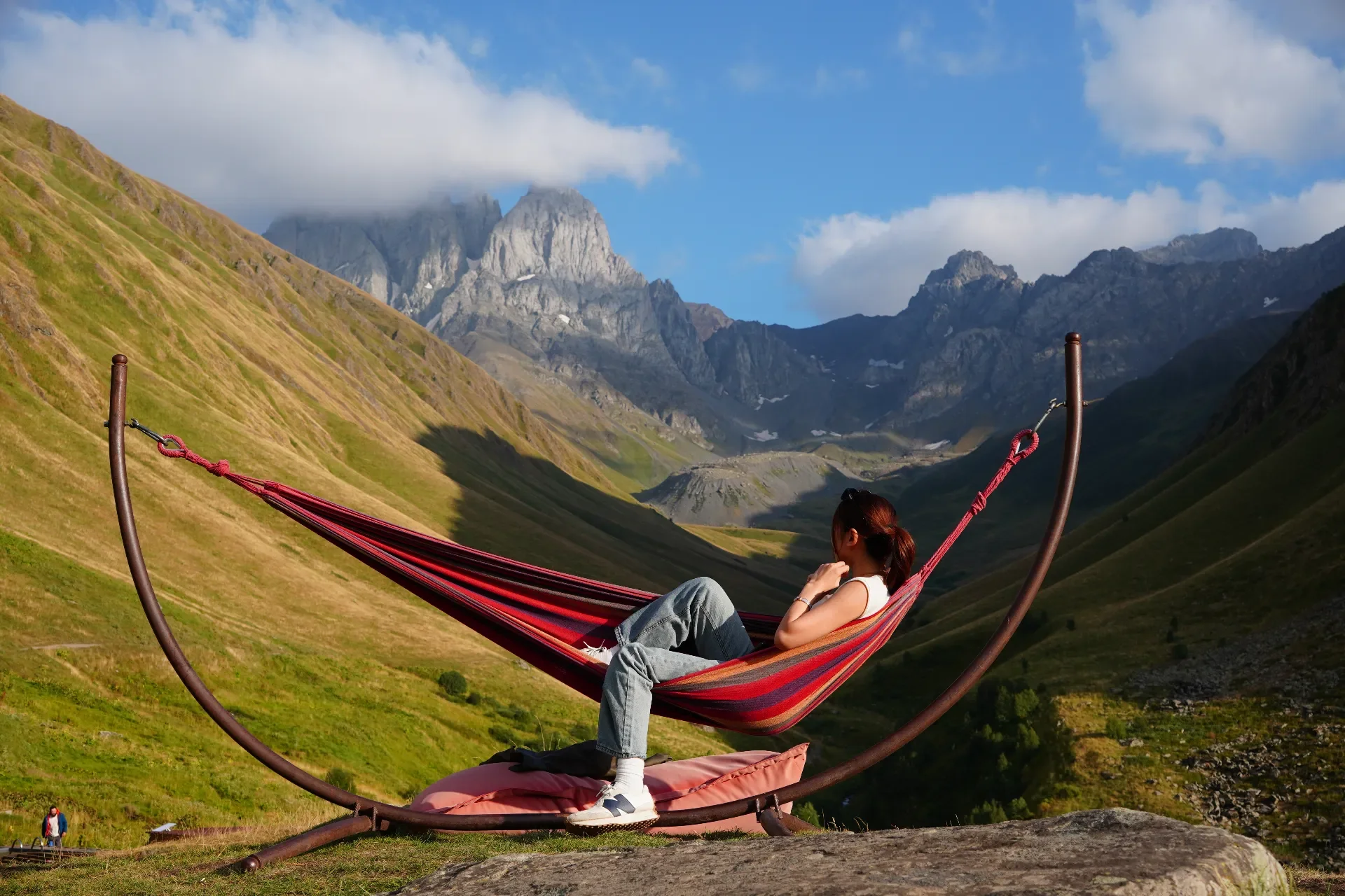 A woman relaxing on a hammock in a mountainous landscape with green hills, rocky cliffs, and cloudy sky.