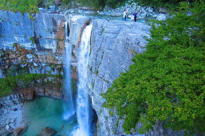 A tall waterfall flowing into a turquoise pool, surrounded by rocky cliffs and green foliage, with three people standing at the top of the cliff.