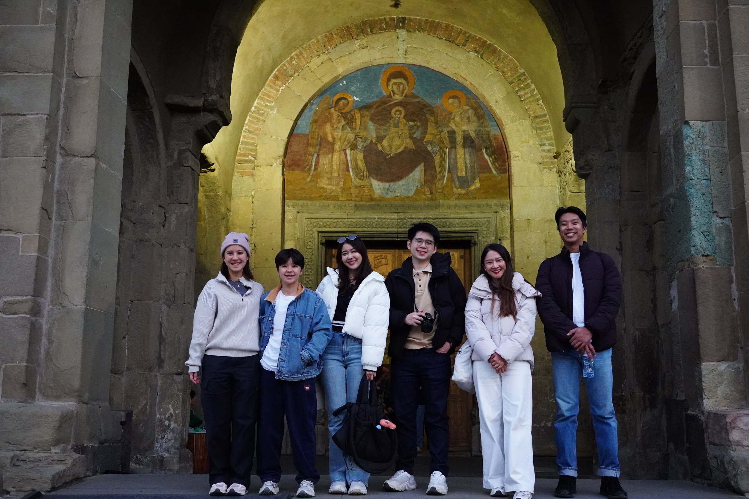 Six young adults standing together outdoor in front of a historical building with religious artwork, smiling for a group photo.