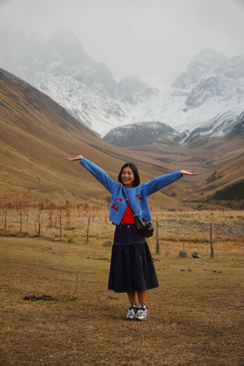 A woman smiling with arms outstretched in a mountainous landscape with snow-capped peaks and brown grass.