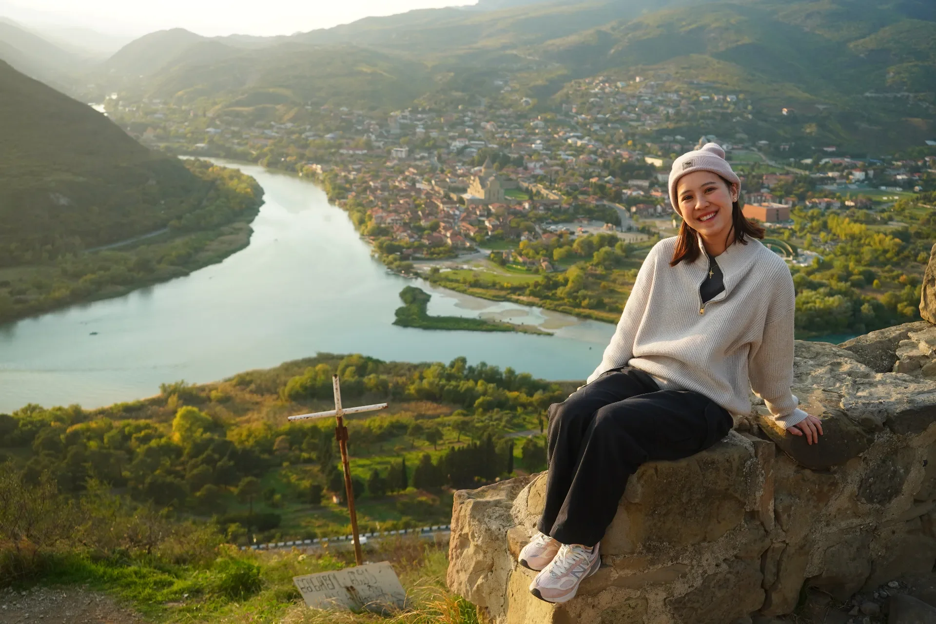 Smiling young woman sitting on a stone wall overlooking a river and town nestled in green hills during sunset.