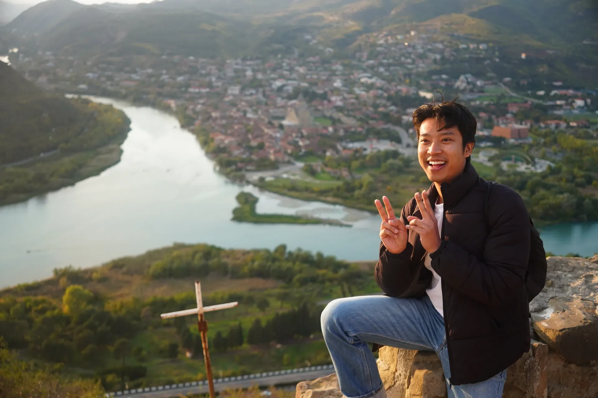 Smiling young man in black jacket and jeans sitting on a rock formation making a peace sign with both hands, overlooking a river and town in a valley with green hills in the background during sunset.