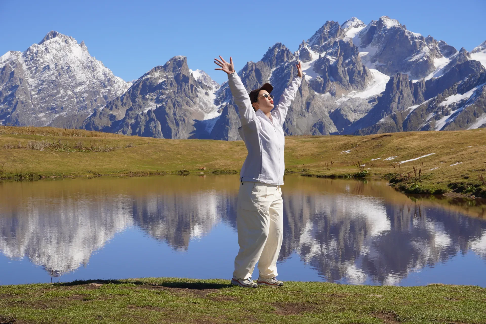 A woman standing with her arms raised in front of a mountain landscape with snow-capped peaks and a lake reflecting the mountains.