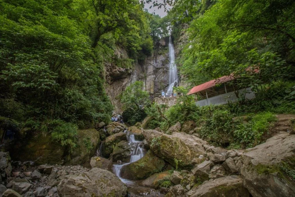 A lush green forest with a waterfall cascading down a rocky cliff into a small stream surrounded by large rocks, with a reddish roof shelter on the right side.