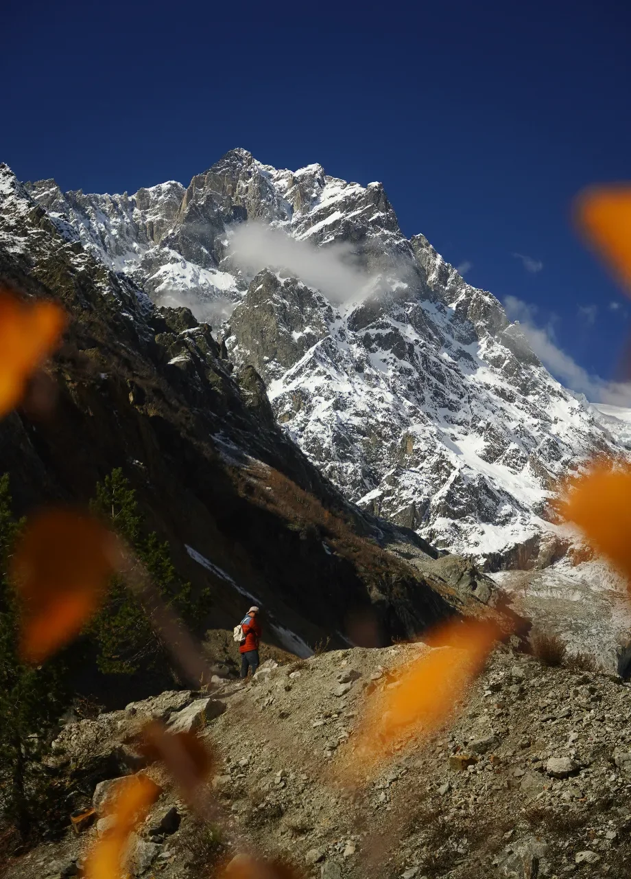 A hiker in a red jacket and white hat walking on a rocky trail in a mountainous landscape with snow-capped peaks and a clear blue sky.