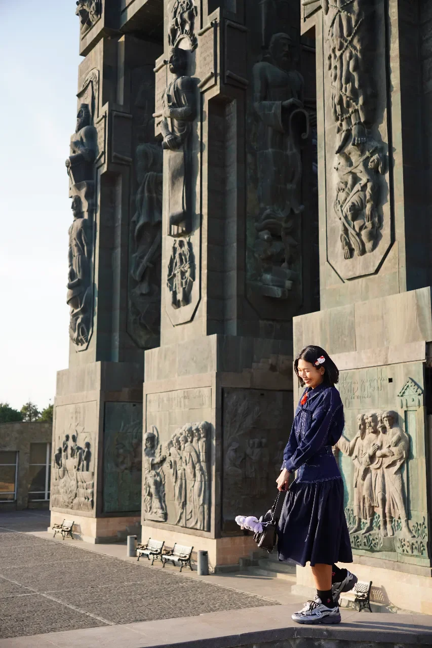 A young woman standing in front of a large stone monument with detailed bas-relief sculptures depicting historical scenes. She is smiling, holding a small bag, dressed in a navy jacket and a black skirt, and wearing sneakers.