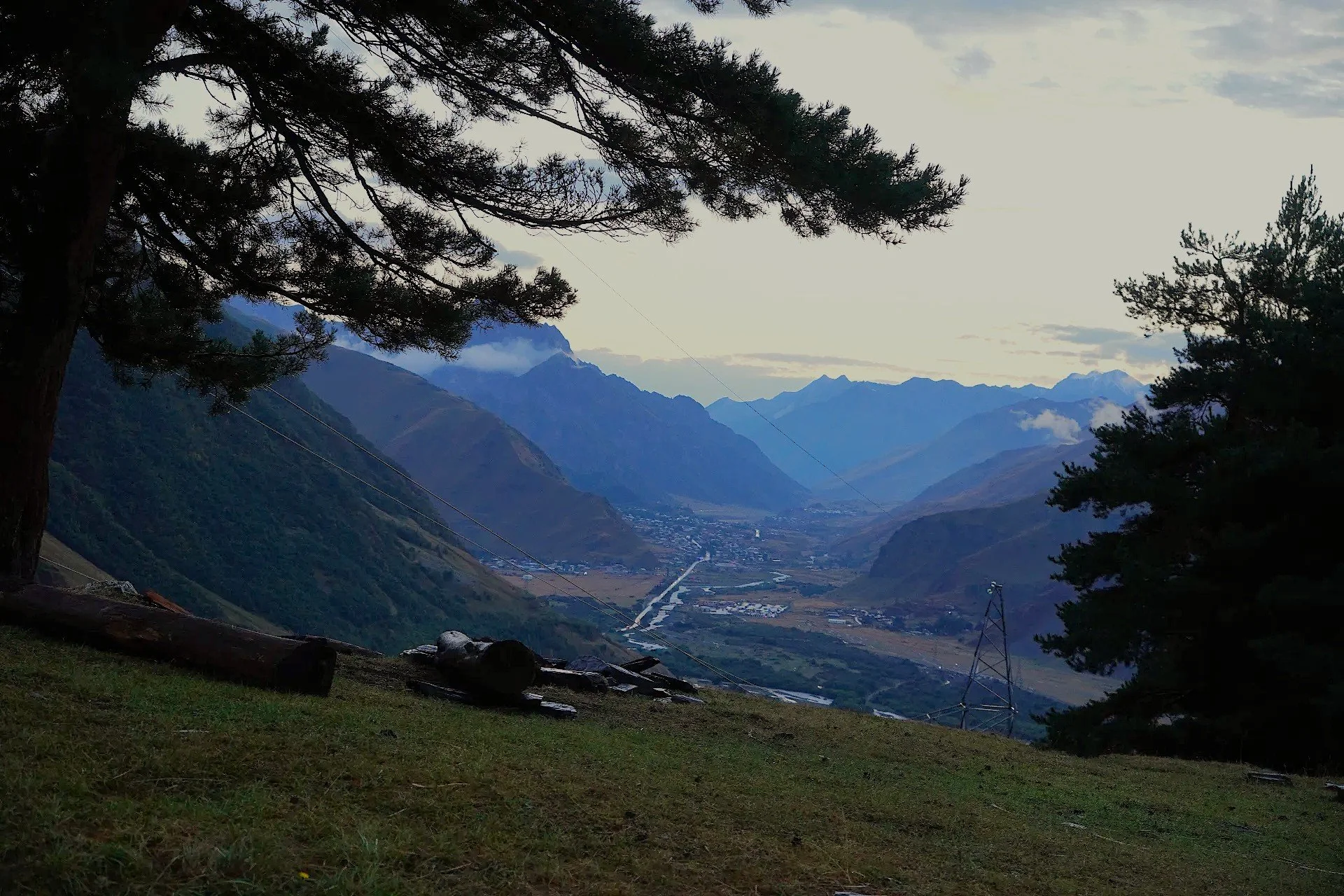 Scenic mountain landscape at dusk with layers of mountain ranges, cloudy sky, and some trees in the foreground.
