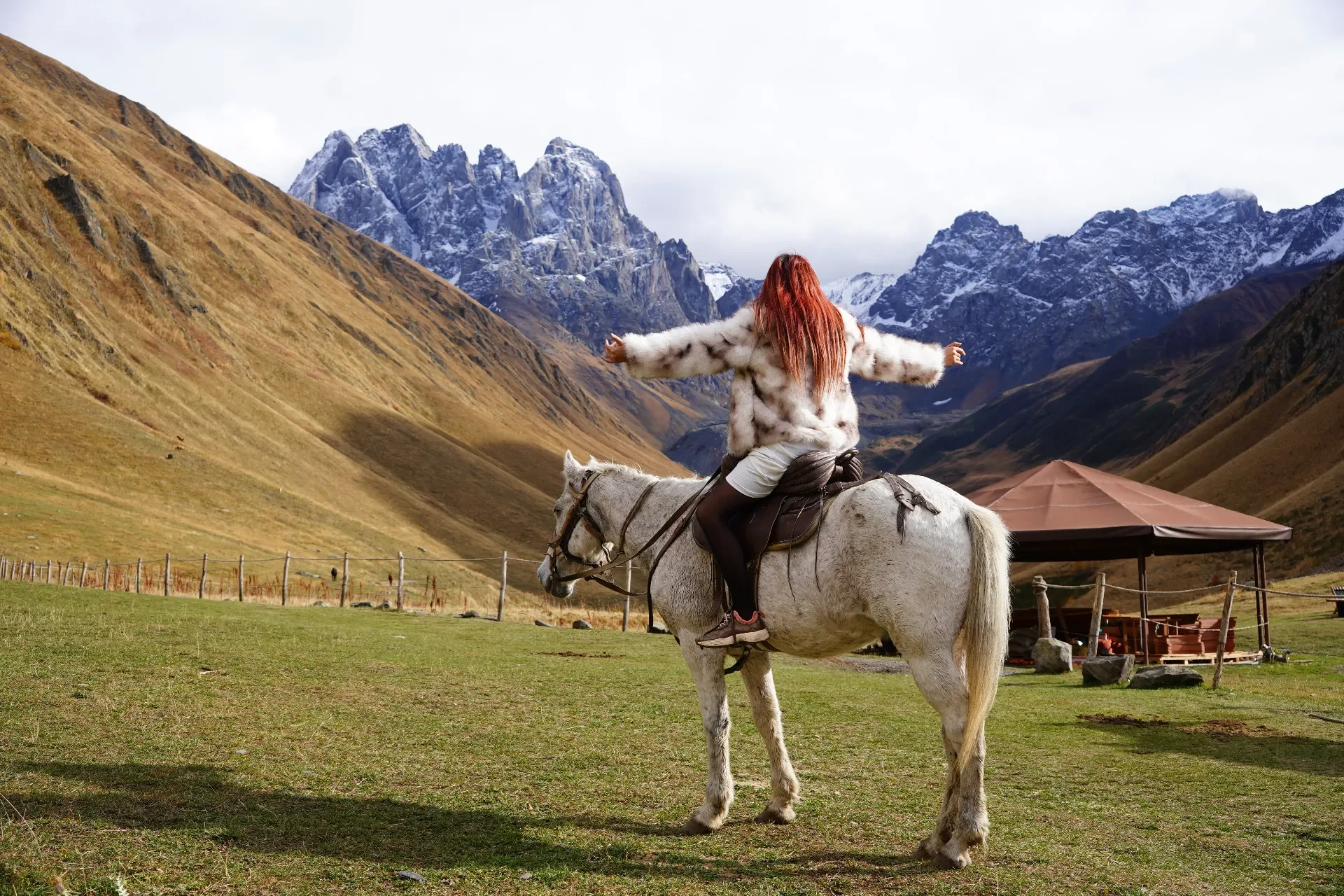 A woman with red hair and a fur coat riding a white horse in a mountain valley with snow-capped peaks in the background.