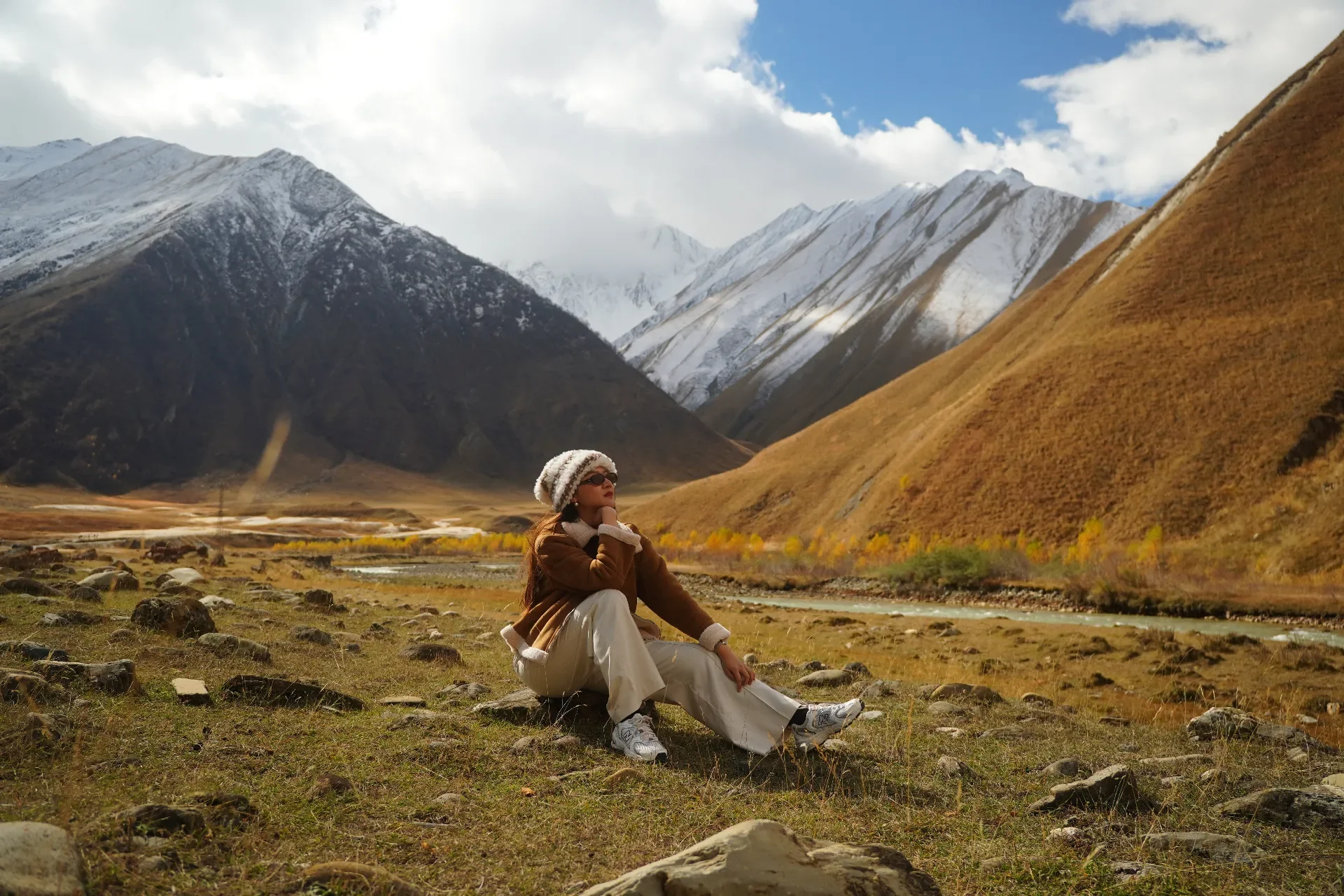 A woman sitting on the grass in a mountain valley with snow-capped peaks in the background, wearing a knit hat, sunglasses, and warm clothing.