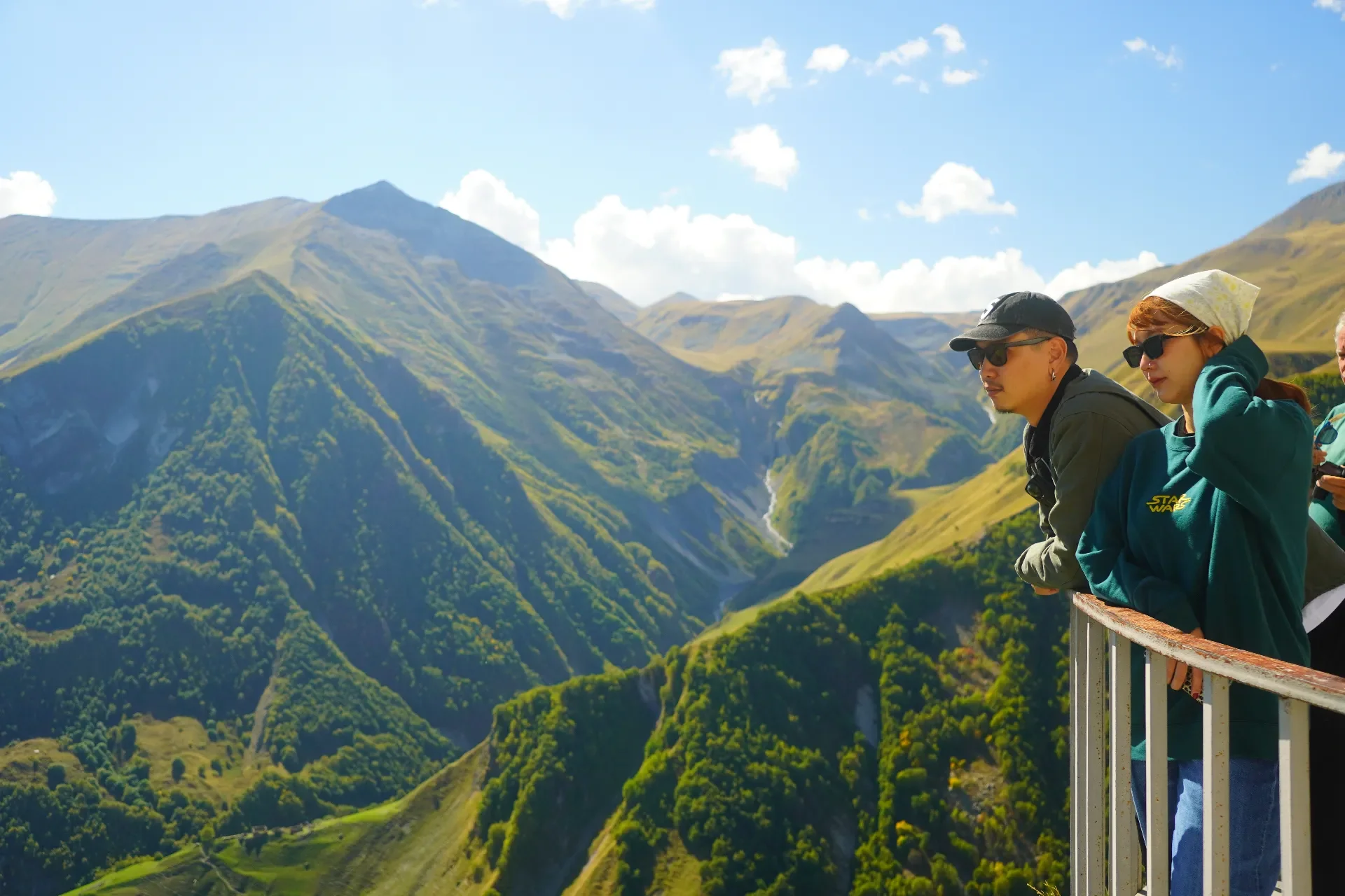 Two people standing on a balcony overlooking green mountains under a partly cloudy sky.