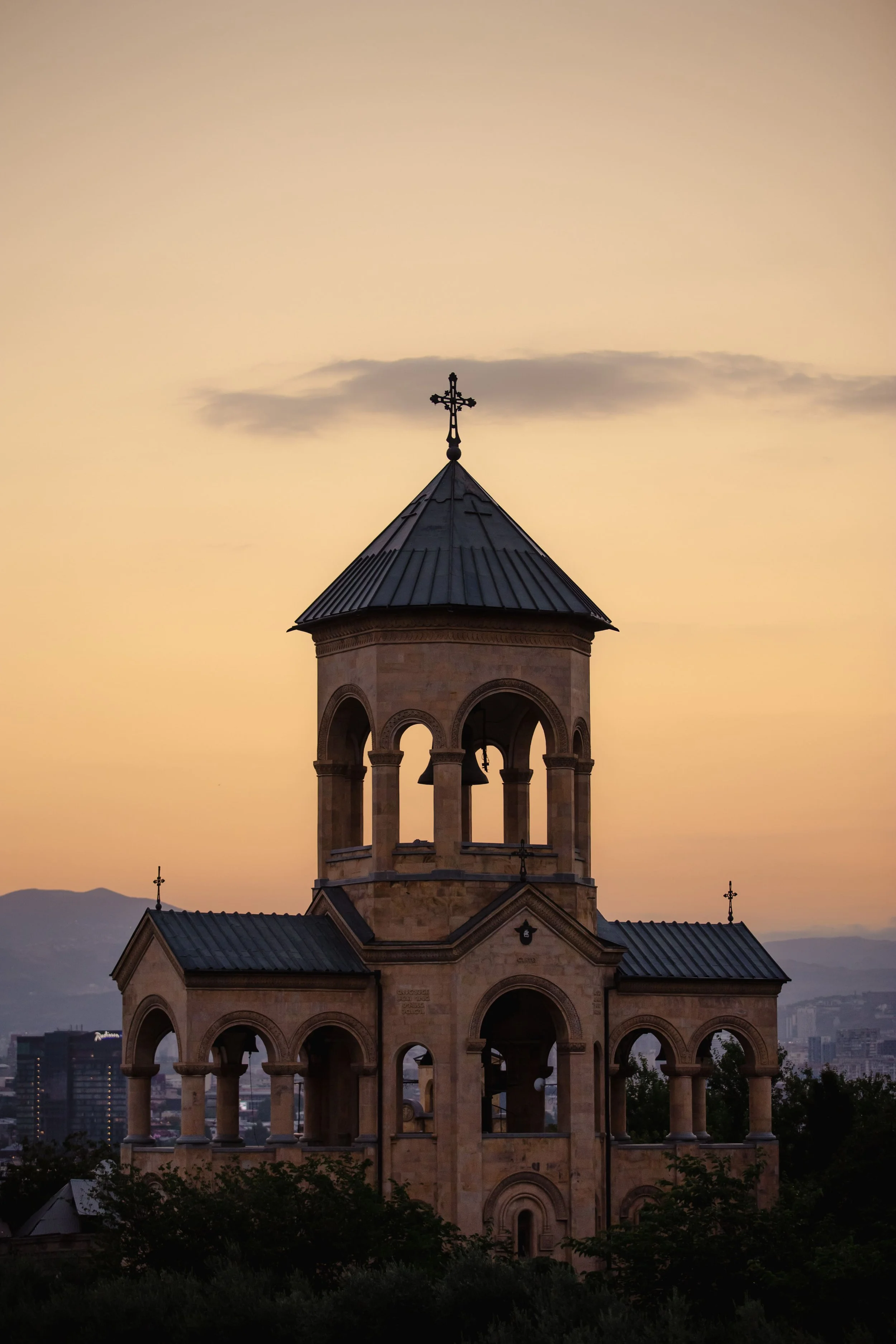 A church tower with a cross on top at sunset, with a cityscape and mountains in the background.