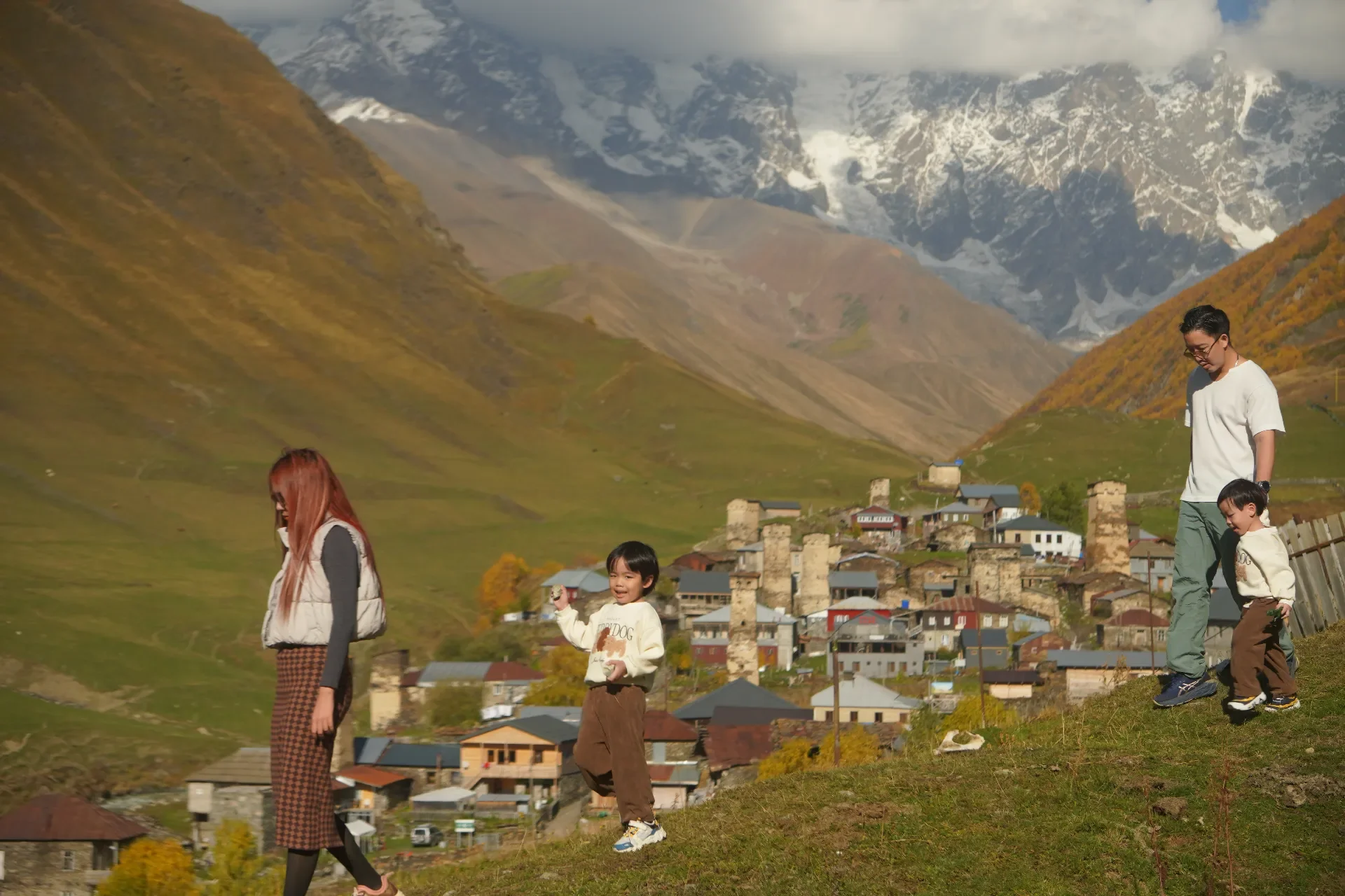 A woman and three children exploring a hillside overlooking a small mountain village with colorful houses and stone towers, surrounded by green and fall-colored trees, with snow-capped mountains in the background.
